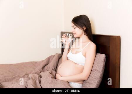 Foto von ziemlich schwangeren Frau halten ihre Hände auf dem Bauch, während Trinkwasser auf dem Bett zu Hause. Stockfoto