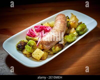 Ungarische Kolbasz-Wurst mit Rosenkohl, Kartoffeln und eingelegtem Kohl auf weißem Teller, Weitblick Stockfoto