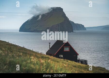 Schwarzes Haus auf dem berühmten faroese Witches Finger Trail und der Insel Koltur im Hintergrund. Sandavagur Dorf, Vagar Insel, Färöer Inseln, Dänemark Stockfoto