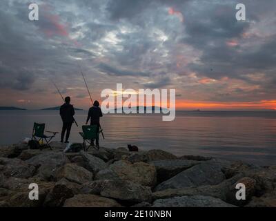 Zwei Fischer mit Stühlen und Ruten stehen während des Sonnenuntergangs auf Felsen und versuchen, einige Fische zu fangen. Eine Katze liegt neben ihnen und wartet auf Nahrung. Stockfoto