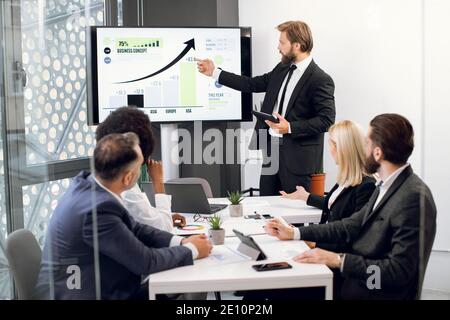 Vorderansicht des jungen selbstbewussten bärtigen Geschäftsmannes, der statistische Daten auf dem großen Plasmabildschirm für seine fokussierten hochqualifizierten multiethnischen Kollegen zeigt Stockfoto