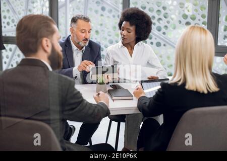 Geschäftsleute aus verschiedenen Rassen, die sich im Sitzungssaal des Büros treffen. Bärtiger älterer Mann, mit Tablet-pc, diskutieren Geschäftsstrategie mit seinem charmanten Stockfoto