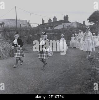 1950, historisch, zwei junge Jungen in Kilts führen die Rose Queen und ihre Gefolge von jungen Mädchen in ihren Kleidern durch einen lokalen Park, zu dem die Zeremonie stattfinden wird, Farnworth, Lancashire, England, Großbritannien. Der Tag der Rosenkönigin war ein besonderer Tag für die Kinder der Stadt, die an der Sonntagsschule der Kirche teilnahmen und mit der traditionellen "Sommersonnenwende" verbunden waren, die ihren Ursprung in den alten heidnischen Festen hatte. Es wurde manchmal zusammen mit dem Walking Day getan, wenn die Kinder versammelten sich und gingen durch die Straßen. Stockfoto