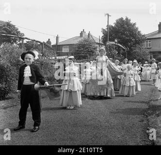 1950, historisch, ein kleiner Junge, der als Stadtschreier gekleidet ist und die Rose Queen und ihr Gefolge kleiner Mädchen in ihren Kleidern auf einem Pfad in einem lokalen Park in Farnworth, Lancashire, England, Großbritannien, führt. Der Tag der Rosenkönigin war ein besonderer Tag für die Kinder der Stadt, die an der Sonntagsschule der Kirche teilnahmen und mit der traditionellen "Sommersonnenwende" verbunden waren, die ihren Ursprung in den alten heidnischen Festen hatte. Es wurde manchmal zusammen mit dem Walking Day getan, wenn die Kinder versammelten sich und gingen durch die Straßen. Stockfoto