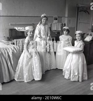 1950s, historisch, eine junge Rose Queen in ihrem Kleid mit ihrem kleinen weiblichen Gefolge gekleidet, eine hält das Kissen für ihre Krone, zusammen in der Dorfhalle nach der Zeremonie und Krönung, Farrnworth, Lancashire, England, Großbritannien. Die Krönung der Rosenkönigin war ein besonderer Tag für die Kinder der Stadt, die die Sonntagsschule der Kirche besuchten und mit der traditionellen "Sommersonnenwende" verbunden waren, die ihren Ursprung in den alten heidnischen Festen hatte. Es wurde manchmal zusammen mit dem Walking Day getan, wenn die Kinder versammelten sich und gingen durch die Straßen. Stockfoto