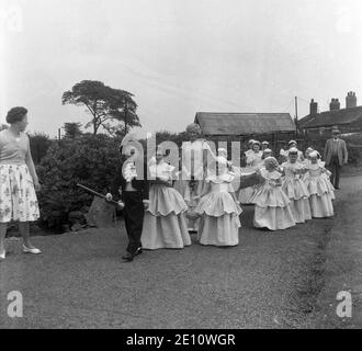 1950s, historisch, ein kleiner Junge in einem Kostüm als Stadt Krier führt die Rose Queen und ihre Gefolge von kleinen Mädchen auf einem Weg in einem lokalen Park in Farnworth, Lancashire, England, Großbritannien. Der Tag der Rosenkönigin war ein besonderer Tag für die Kinder der Stadt, die an der Sonntagsschule der Kirche teilnahmen und mit der traditionellen "Sommersonnenwende" verbunden waren, die ihren Ursprung in den alten heidnischen Festen hatte. Es wurde manchmal zusammen mit dem Walking Day getan, wenn die Kinder versammelten sich und gingen durch die Straßen. Stockfoto