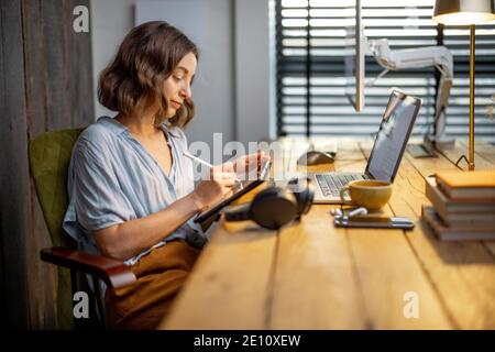 Junge Frau, die lässig gekleidet ist und kreative Arbeit hat, auf einem digitalen Tablet zeichnet und im gemütlichen und stilvollen Home Office sitzt Stockfoto