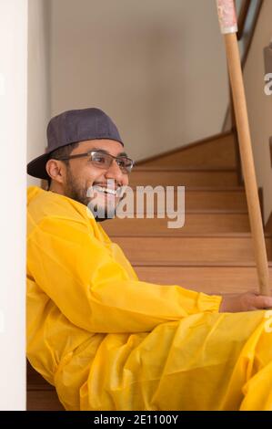 Arbeiter Mann in gelber Regenkleidung sitzt auf dem Rand von einigen hölzernen Tribüne mit Kappe, Brille Bart und Lächeln an einem hellen Tag Stockfoto