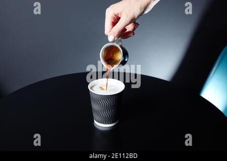 Der Barista gießt Espresso in die Einweg-Papiertasse auf dem Holztisch im Café. Zubereitung von leckeren Kaffee trinken mit Milch Stockfoto
