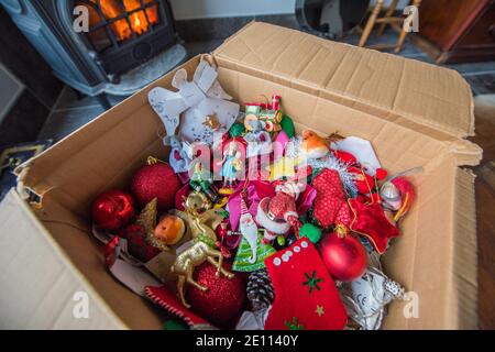 Braune Pappschachtel mit einem Durcheinander von Familie bunte Weihnachten Baumschmuck vor einem Holzbrenner auf einem Stein Boden wird weggepackt Stockfoto