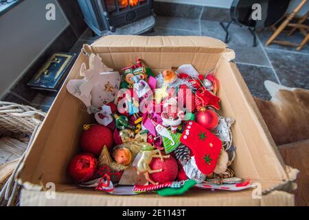 Braune Pappschachtel mit einem Durcheinander von Familie bunte Weihnachten Baumschmuck vor einem Holzbrenner auf einem Stein Boden wird weggepackt Stockfoto