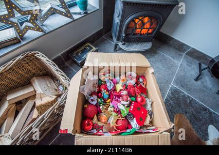 Braune Pappschachtel mit einem Durcheinander von Familie bunte Weihnachten Baumschmuck vor einem Holzbrenner auf einem Stein Boden wird weggepackt Stockfoto