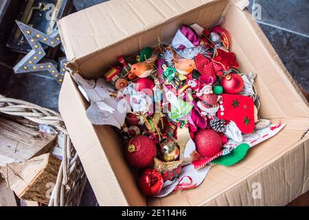 Braune Pappschachtel mit einem Durcheinander von Familie bunte Weihnachten Baumschmuck vor einem Holzbrenner auf einem Stein Boden wird weggepackt Stockfoto