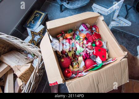 Braune Pappschachtel mit einem Durcheinander von Familie bunte Weihnachten Baumschmuck vor einem Holzbrenner auf einem Stein Boden wird weggepackt Stockfoto