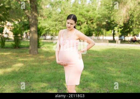 Eine schwangere junge Frau im Park und hält sich an den Magen, der schmerzt. Schwangerschaft und Mutterschaft. Rückenschmerzen während der Schwangerschaft. Stockfoto