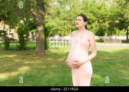 Eine schwangere junge Frau im Park und hält sich an den Magen, der schmerzt. Schwangerschaft und Mutterschaft. Rückenschmerzen während der Schwangerschaft. Stockfoto