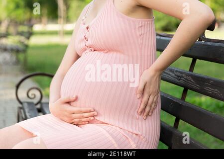 Eine schwangere junge Frau auf einer Bank sitzt im Park und hält sich an den Bauch, der schmerzt. Schwangerschaft und Mutterschaft. Rückenschmerzen während der Schwangerschaft. Stockfoto