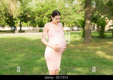 Eine schwangere junge Frau im Park und hält sich an den Magen, der schmerzt. Schwangerschaft und Mutterschaft. Rückenschmerzen während der Schwangerschaft. Stockfoto