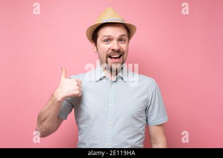 Attraktive fröhliche junge Mann im Sommer Hut zeigt Daumen nach oben. Studio auf gelber Wand. Stockfoto