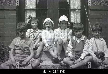 Circa 1940er Jahre, ein charmantes historisches Bild von drei kleinen Jungen und drei kleinen Mädchen, die vor einem Paar französischer Gartentüren für ein Foto sitzen. Es sieht so aus, als ob die Jungen ein Spiel mit Bögen und Pfeilen gespielt haben, wobei ein Junge, der größte, einen Bogen um seine Taille hat, während die anderen beiden Jungen Pfeile halten. Der Junge auf der linken Seite trägt einen Fleck über seinem linken Auge, vielleicht ein Unfall von Stunden von gesunden Outdoor-Spaß für die Jungs zur Verfügung gestellt. Stockfoto