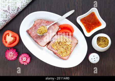 Geröstetes Schweinefleisch auf einem Teller mit französischem Senf, Tomaten, Chilisauce und Blumen. Selektiver Fokus Stockfoto