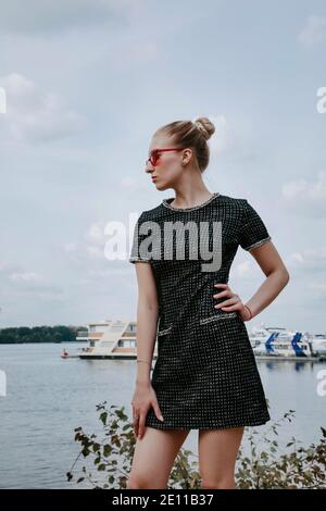 Das hübsche blonde Mädchen steht am Wasser mit Booten im Hintergrund; sie trägt ein stilvolles kurzes dunkles Kleid und eine rote Sonnenbrille. Stockfoto