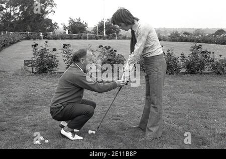 70er Jahre, historisch, draußen auf einem Grasrand von einem Putting Green, ein älterer männlicher Golfprofi zeigt einen jungen Amateur männlichen Golfspieler, der den Golfclub hält, der "Vardon Grip", auch bekannt als der überlappende Griff, benannt nach dem langen Golfer Harry Vardon, einem sechsfachen Open Champion. Um das Golfspiel gut zu spielen, ist der richtige Griff wichtig. Beachten Sie die Kleidung des langhaarigen jungen Mannes, der ein Hemd und eine kipper Krawatte und Flair Hose der Zeit trägt. Stockfoto