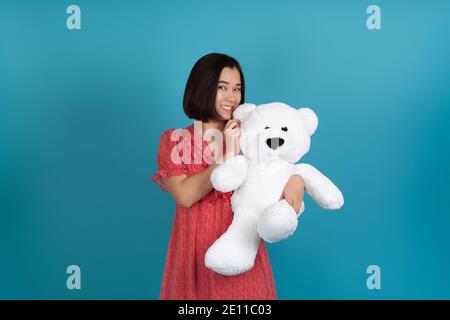 Nahaufnahme Porträt einer schlauen, schelmischen jungen lächelnden Frau in einem roten Kleid mit einem weißen Teddybär am Ohr, isoliert auf einem blauen Hintergrund Stockfoto