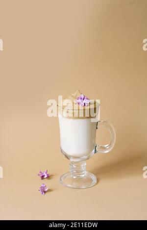 Schöner Dalgon-Kaffeedrink mit Schaum in einer transparenten Tasse und lila Blüten auf beigem Hintergrund. Speicherplatz kopieren Stockfoto
