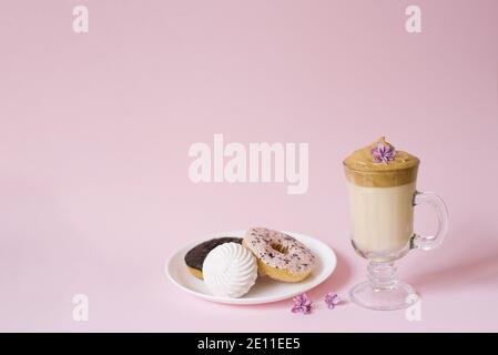 Schöner Dalgon-Kaffee-Drink. Eine Tasse Kaffee mit Schaum und Flieder Blume, ein Teller mit Süßigkeiten: Marshmallows und Donuts auf einem Flieder Hintergrund. Kopieren von n Stockfoto