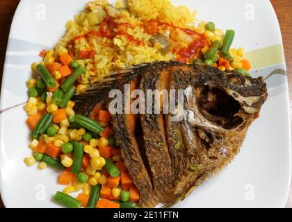 Gebratene Tilapia mit gelbem Reis, heißer Sauce und gemischtem Gemüse auf einem weißen Teller Stockfoto