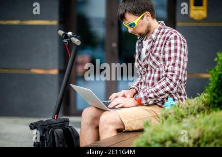 Attraktive junge männliche Freiberufler in Sonnenbrille hält einen Laptop und schaut sorgfältig auf Monitor, sitzt Bank draußen, nutzt Elektroroller, lässigen Stil Stockfoto