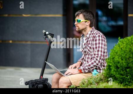 Attraktive junge männliche Freiberufler in Sonnenbrille hält einen Laptop und schaut sorgfältig auf Monitor, sitzt Bank draußen, nutzt Elektroroller, lässigen Stil Stockfoto