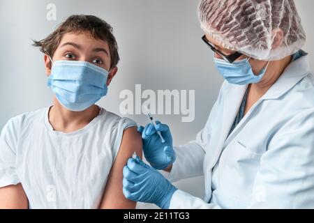 Ängstlich Kind macht Gesicht, Angst vor der Spritze. Medic, Arzt, Krankenschwester, Heilpraktiker in weißem Kleid und Gesichtsmaske impft Teenager. Stockfoto