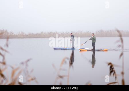 Zwei Männer paddeln an einem windstillen Wintermorgen auf einem ruhigen Fluss, umgeben von Nebel, ruhigem Wasser und Schilf Stockfoto