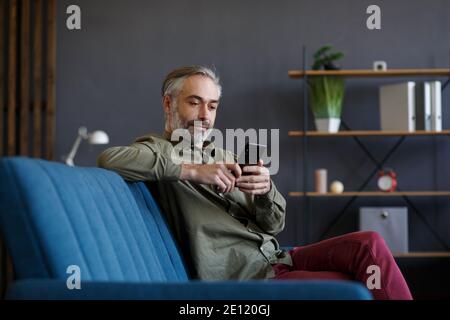 Lächelnder grauhaariger Mann mit Handy-Apps, SMS-Nachrichten, Surfen im Internet, Online-Shopping. Blick auf Smartphone und lächelnd, sitzen an Stockfoto