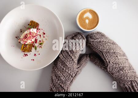 Brauner Fäustling und Becher mit Cappuccino und Kuchen auf weißem Holztisch. Weihnachten und Neujahr Hintergrund. Draufsicht, Kopierbereich. Kuschelige Komposition. Winter Stockfoto