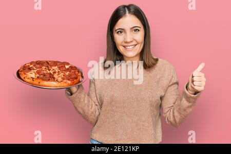 Junge schöne Frau hält italienische Pizza lächelnd glücklich und positiv, Daumen nach oben tun ausgezeichnet und Zustimmung Zeichen Stockfoto