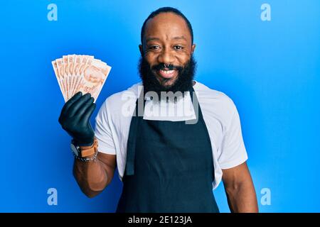 Junger afroamerikanischer Mann, der eine professionelle Schürze mit türkischen Lira trägt Banknoten, die positiv und glücklich stehen und lächeln mit einem Selbstbewusst Stockfoto