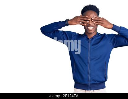 Junger afroamerikanischer Mann trägt Sportkleidung, die Augen mit lächelnden Händen bedeckt, fröhlich und lustig. Blind-Konzept. Stockfoto