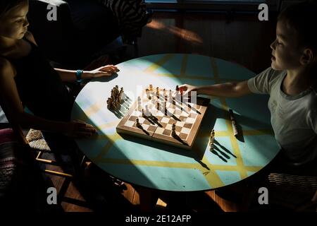 Ein junges Mädchen sieht zu, wie ihr kleiner Bruder ein Stück bewegt, während sie zusammen Schach spielt und an einem niedrigen Tisch sitzt, der im Sonnenlicht gebadet ist. Stockfoto