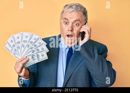 Senior grauhaarige Mann trägt Anzug hält Dollar und mit Smartthone ahnungslos und verwirrt Ausdruck. Zweifel Konzept. Stockfoto