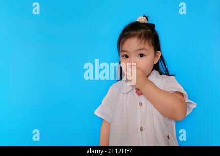 Ein süßes junges asiatisches Mädchen versucht, ein Geheimnis zu bewahren, bedeckt ihren Mund mit ihrer Hand und verhält sich verdächtig. Einfacher hellblauer Hintergrund. Stockfoto