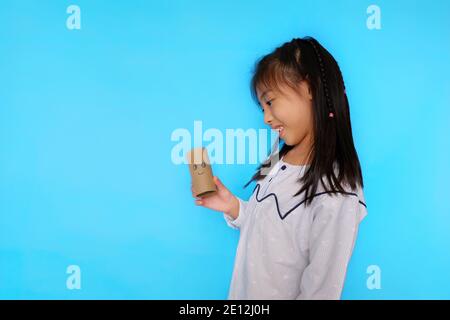 Ein süßes junges asiatisches Mädchen, das mit einer leeren Seidenrolle spielt und ein Gesicht darauf zeichnet. Einfacher hellblauer Hintergrund. Stockfoto