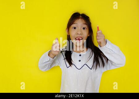 Ein süßes junges asiatisches Mädchen, das einen Haufen Farbstifte hält und sich fröhlich fühlt, nachdem es eine Idee für ihr Kunstprojekt entwickelt hat. Hellgelber Hintergrund. Stockfoto