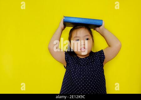 Ein süßes junges asiatisches Mädchen, das versucht, ein großes blaues Textbuch auf ihrem Kopf als Strafe von ihrem Lehrer auszugleichen. Hellgelber Hintergrund. Stockfoto