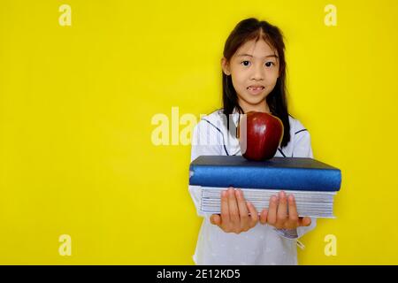 Ein nettes junges asiatisches Mädchen hält einen Stapel von Büchern mit einem roten Apfel auf der Oberseite immer bereit, wieder zur Schule gehen. Hellgelber Hintergrund. Stockfoto