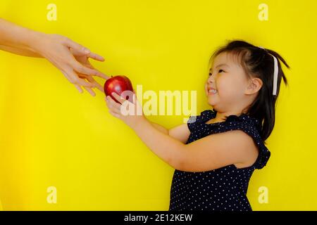 Ein süßes junges asiatisches Mädchen in der Schule, das ihrem Lehrer einen roten Apfel als Geschenk überreicht und lächelt. Hellgelber Hintergrund. Stockfoto