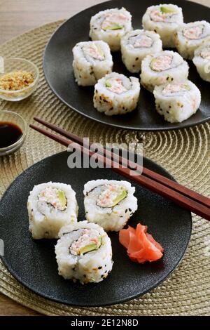 Frisch zubereitete kalifornische Sushi-Rollen auf schwarzen Tellern im vertikalen Format. Rote Ingwerscheiben, Sesamsamen und Sojasauce-Gewürze. Stockfoto