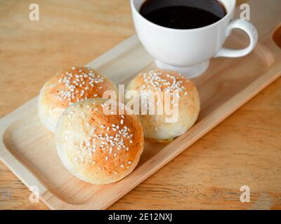 Frische hausgemachte Burger-Brötchen mit weißem Sesam auf einem kleinen Holztablett serviert mit heißem Kaffee auf Holztisch zum Frühstück. Drei leckere süße Sandwiches Stockfoto